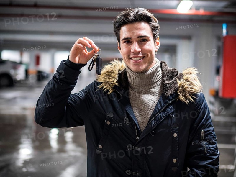 Young trendy man in winter outerwear standing on parking showing alarm remote control to the camera, smiling