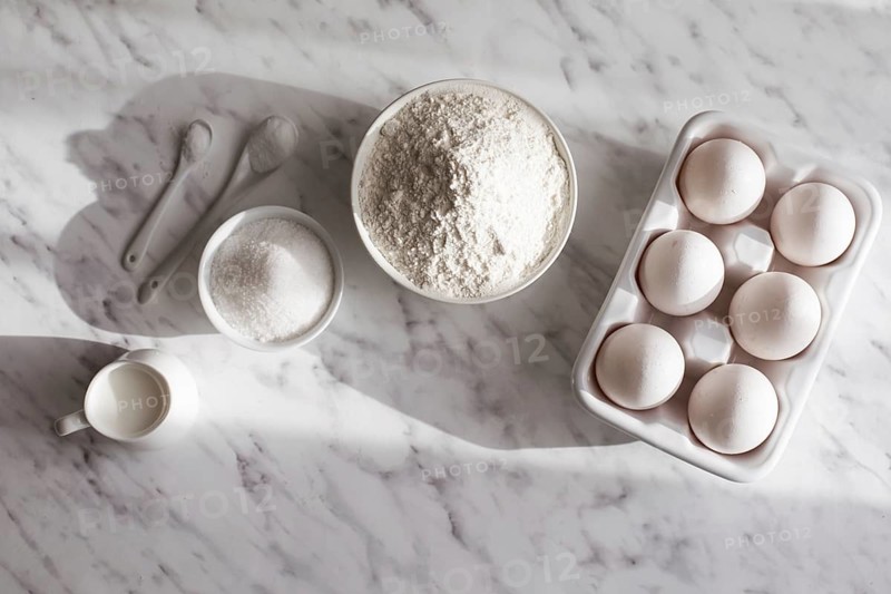 Top view ceramic tray with eggs placed near bowl of flour and sugar. White utensils with white ingredients on white surface. Copy space