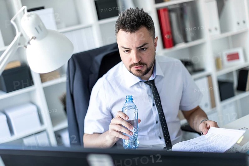 A man is sitting at a table in the office, working with documents and holding a bottle of water in his hand
