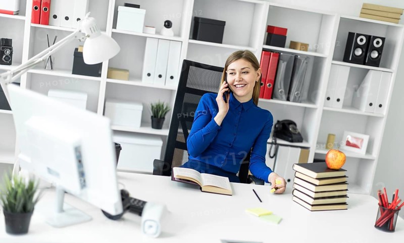 A young girl is sitting at a computer desk, holding a yellow marker in her hand and talking on the phone. Before the girl lies an open book