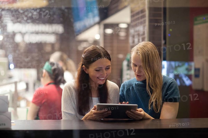 Smiling blonde and brunette women have fun in the restaurant while watching something on the tablet