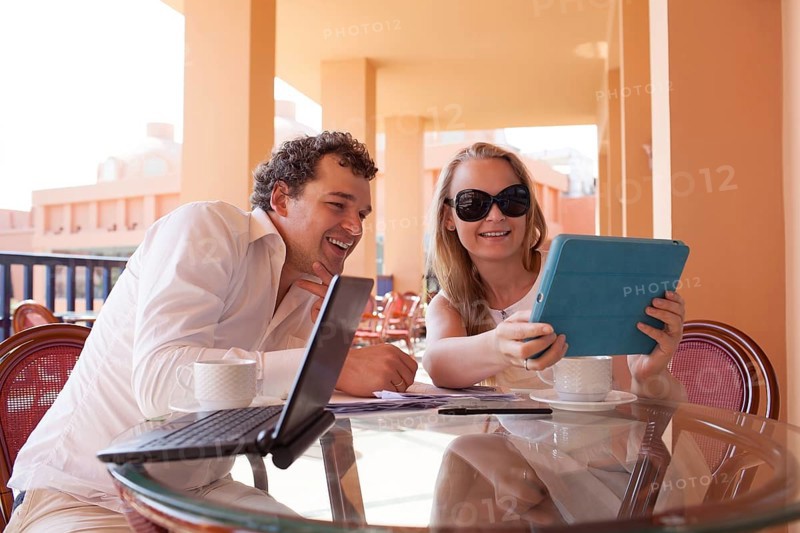 Young couple relaxing over coffee on a balcony sitting at a glass table smiling as they lean close together to read the screen on a tablet computer