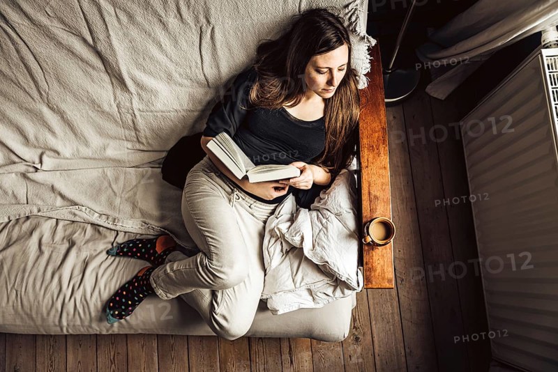 Hig angle view of a 30 yo woman in her sofa, reading a book with a coffee, Brussels