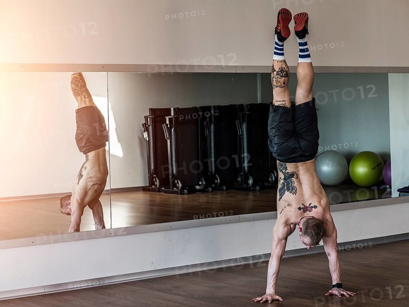 Back view of tattooed shirtless man doing handstand in sportive gym against mirror