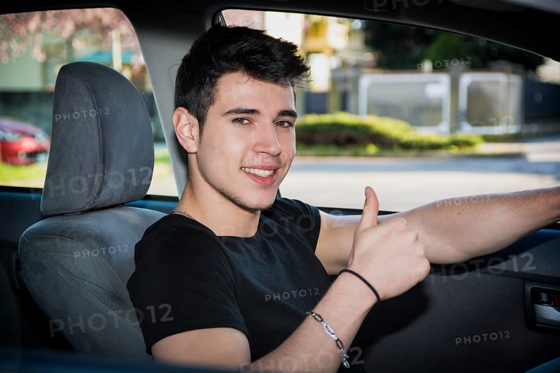 Happy young man or teenager doing thumb up sign while driving car and smiling at camera