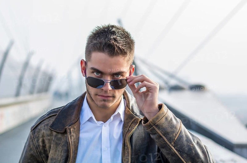 Handsome trendy young man standing on a sidewalk, wearing sunglasses and a fashionable leather jacket, in a relaxed confident pose looking at camera over the glasses