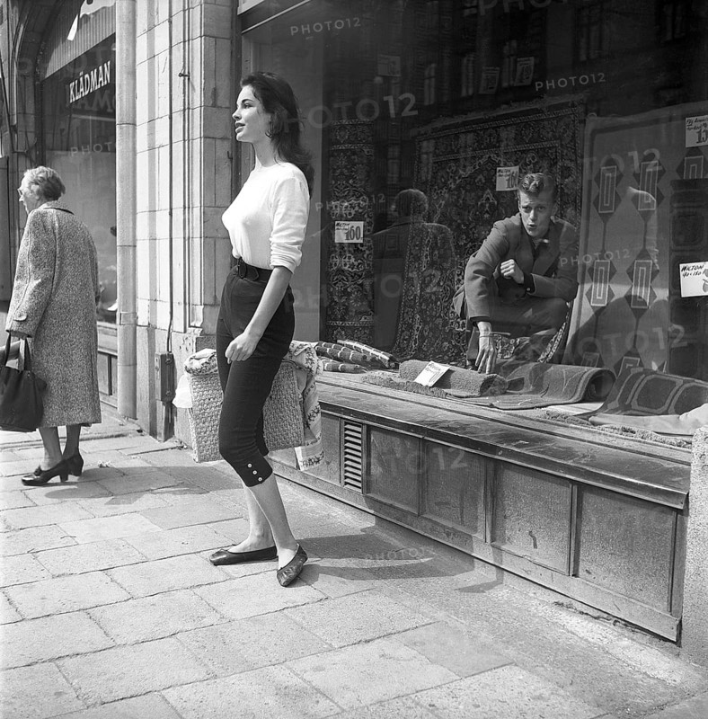 Young woman in 1956. She is standing in front of a shop window on Strandvägen in Stockholm and maybe she is not aware of the guy in the carpet shop who is sitting inside the window and looking at her admiringly. She is wearing a white jumper and knee-high trousers. Sweden 1956. Kristoffersson ref BS94-3
