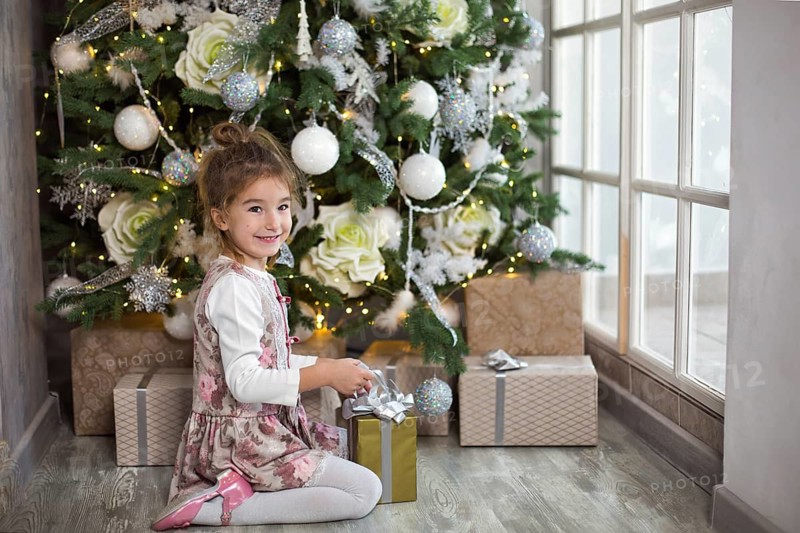 Little girl in beautiful dress is sitting under Christmas tree with gift box and bow. Light from a large window, dark room, home comfort, Christmas, magic and joy. New year, white decor