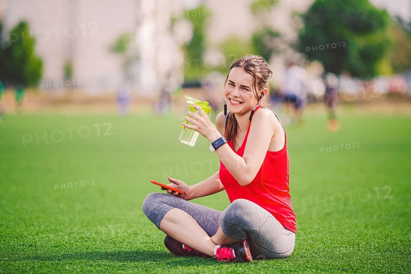 Theme sport health. beautiful young girl sitting resting on green grass lawn stadium using technodogies. In handphone in ear headphones listens music, summer sportswear smart watch heart rate monitor