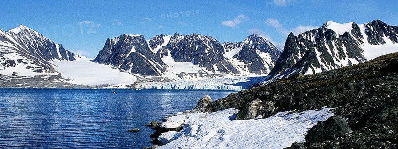 Landscape on the Magdalenenfjord, which is bordered by snow-covered, rugged mountains. At the end of the fjord, glaciers push their ice directly into the dark blue water of the fjord. Bright sunshine, blue sky, deserted. Panorama format. Landscape at Magdalene Fjord, bounded by snow-covered, rugged mountains. At the end of the fjord glaciers push their ice directly into the deep blue waters of the fjord. Panorama view