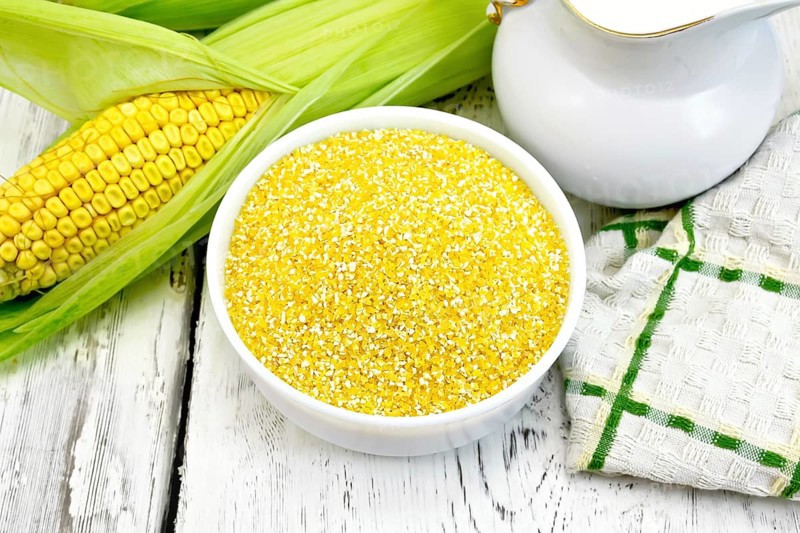 Corn grits in a white bowl, cobs, a jug of milk and a napkin on a wooden boards background