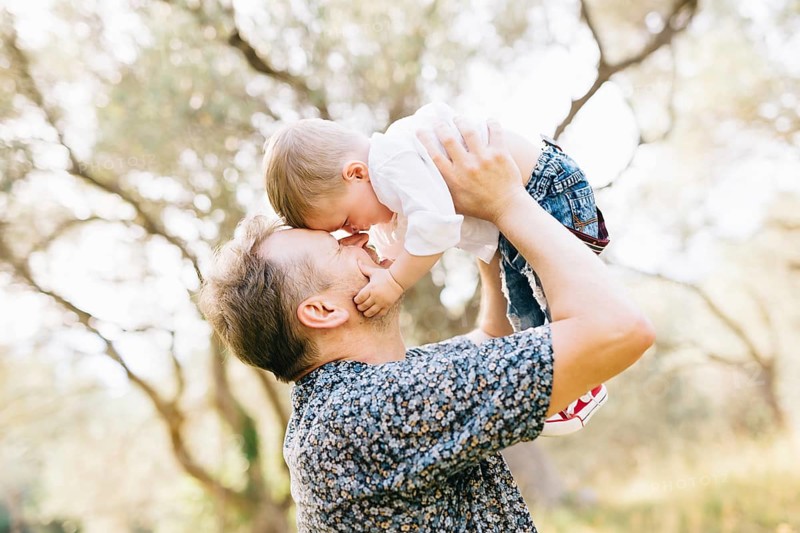 The father holds his son above him, they touching their noses, the son holds the father by the cheek .