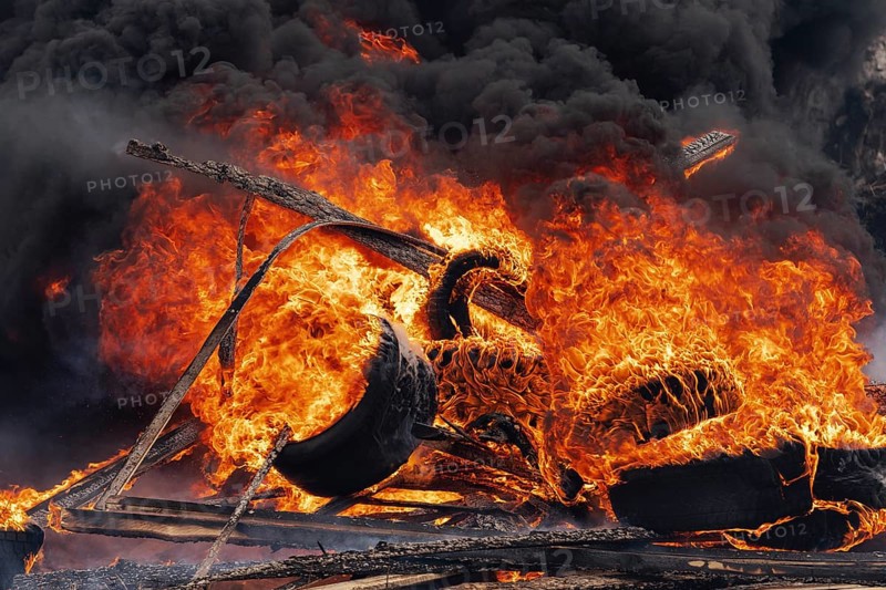 Burning cars wheels, strong flame of red-orange fire and clouds of black fumes in sky. Selective focus, blur from strong fire