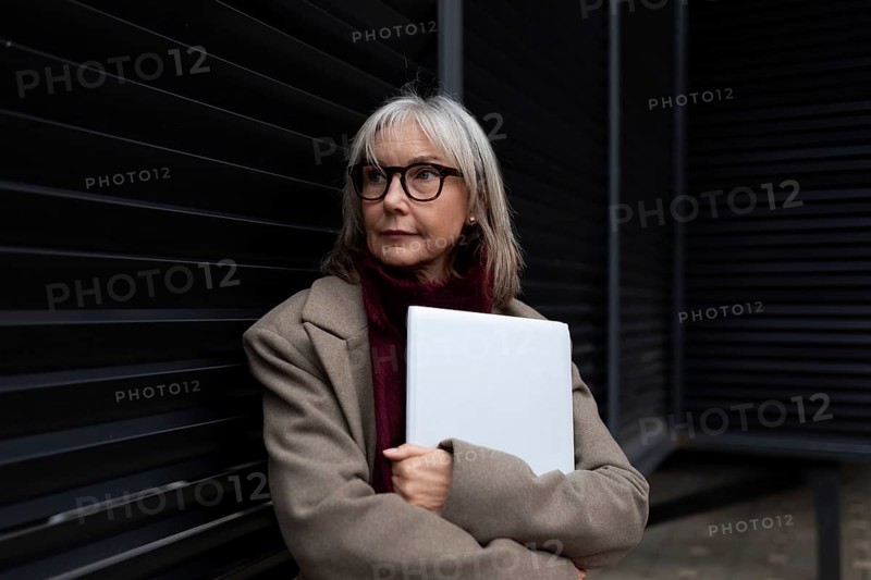 A middle-aged businesswoman with glasses is standing against a dark wall, holding a laptop. She looks thoughtful and confident, dressed in a stylish coat