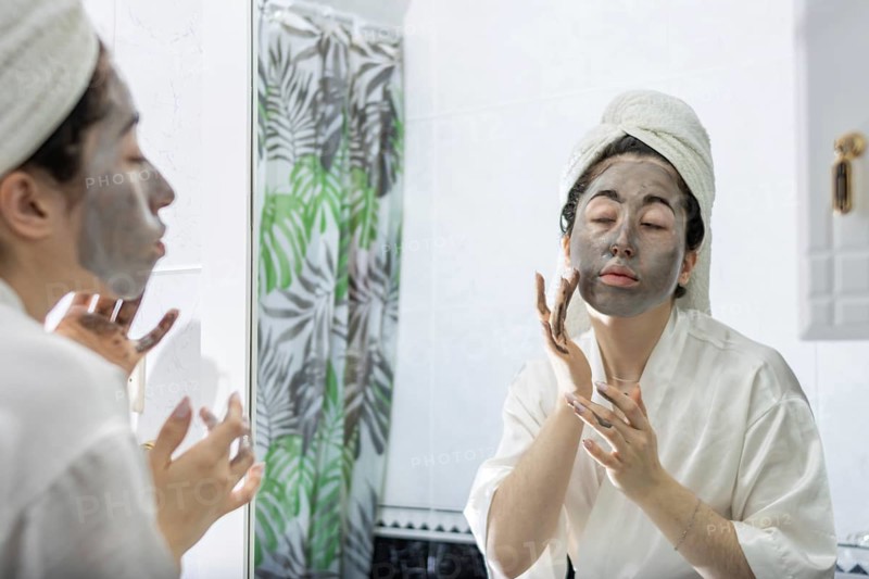 Portrait of a young woman applying a black cream face mask for skin in front of her bathroom mirror after a relaxing shower. Skin care and female beauty. Daily routine