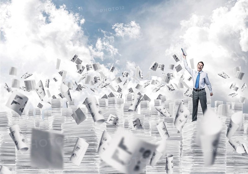 Businessman keeping hand with book up while standing among flying papers with cloudly skyscape on background. Mixed media
