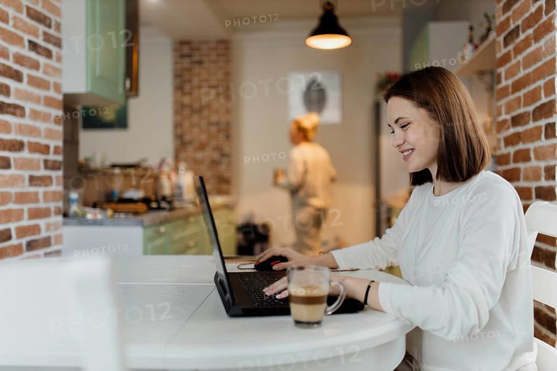 Woman happily using a laptop in a kitchen, enjoying her work with a relaxed demeanor