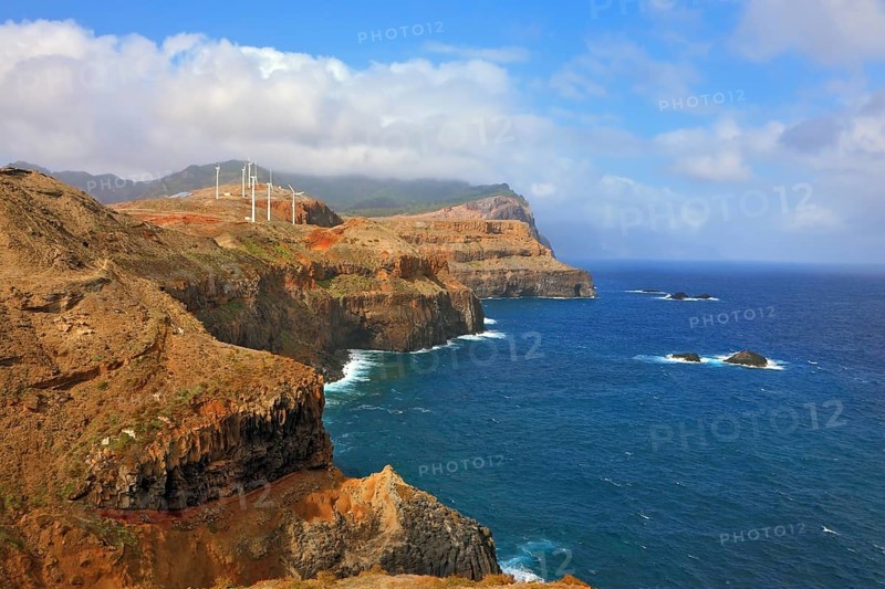 Eastern tip of the island of Madeira. Rocks steeply in the blue waters of the Atlantic Ocean. Over a cliff on the ocean breeze are the windmills