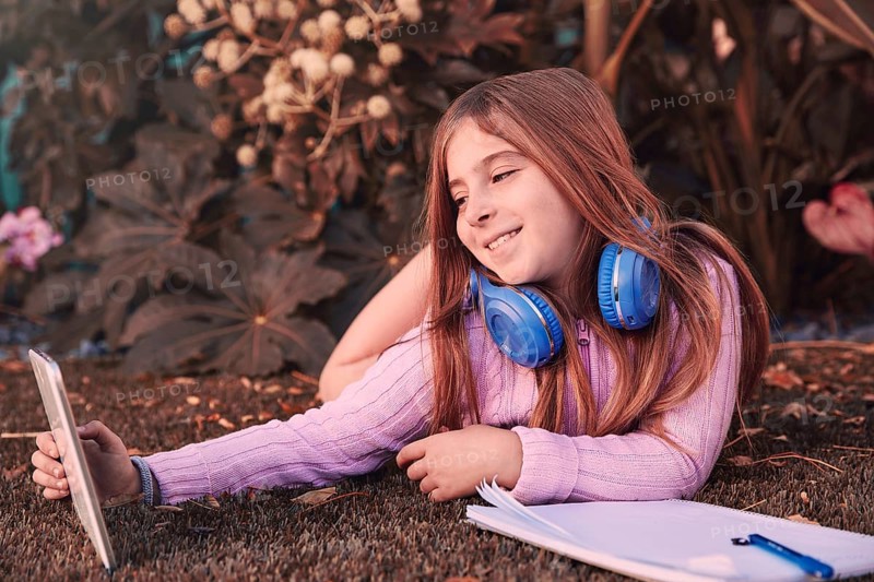 Blond kid girl selfie photo with tablet pc lying on grass turf with headphones