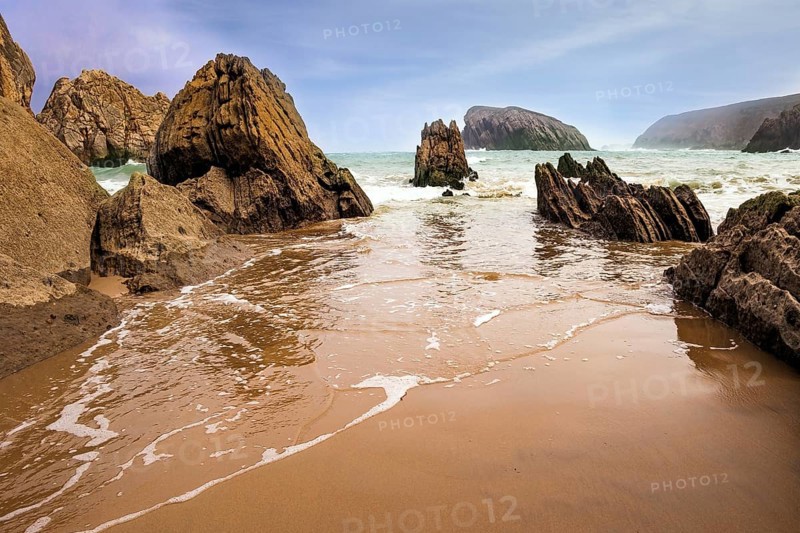 The smooth wet sand of the beach reflects the rocky boulders. The Playa de la Arnia beach. Snow-white sea foam glistens. Spain, Cantabria