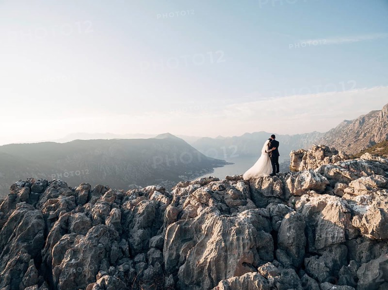Bride and groom are embracing on a rocky mountain and look at the bay. Back view.