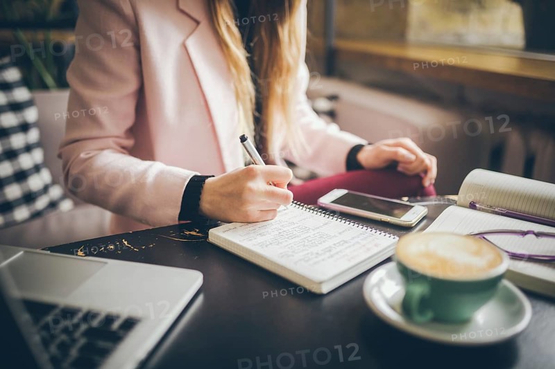 Close up caucasian woman hand on wooden table inside cafe makes notes in notebook. Subject freelancer blogger journalist at work. Unrecognisable person. Technology phone and laptop with cup coffee
