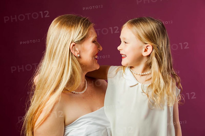 Portrait of happy smiling mother and daughter, both with long loose blond hair, wearing white clothes and red lipstick. Daughter looks like mother. Studio with purple background