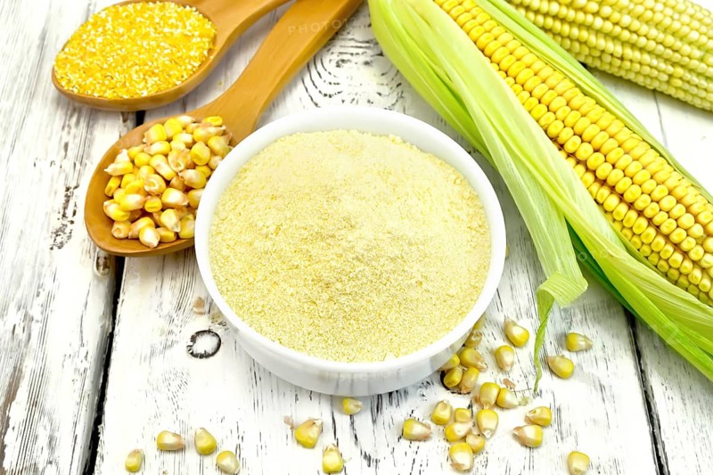 Flour corn in a bowl, groats and grain maize in the spoons, cobs on the background white wooden boards