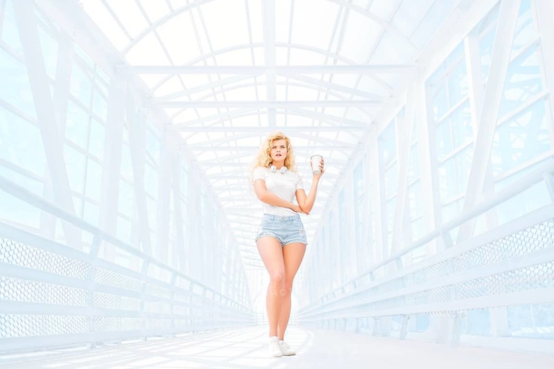 Beautiful young woman with music headphones standing on the bridge with a takeaway coffee mug and posing in front of an urban background