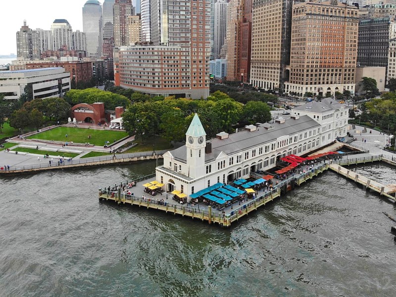 Aerial view of Battery Park pier A leading to Liberty Island, people can be seen waiting in line for boarding as well as looking at Liberty Island located opposite to the docks. October, 22nd, 2019