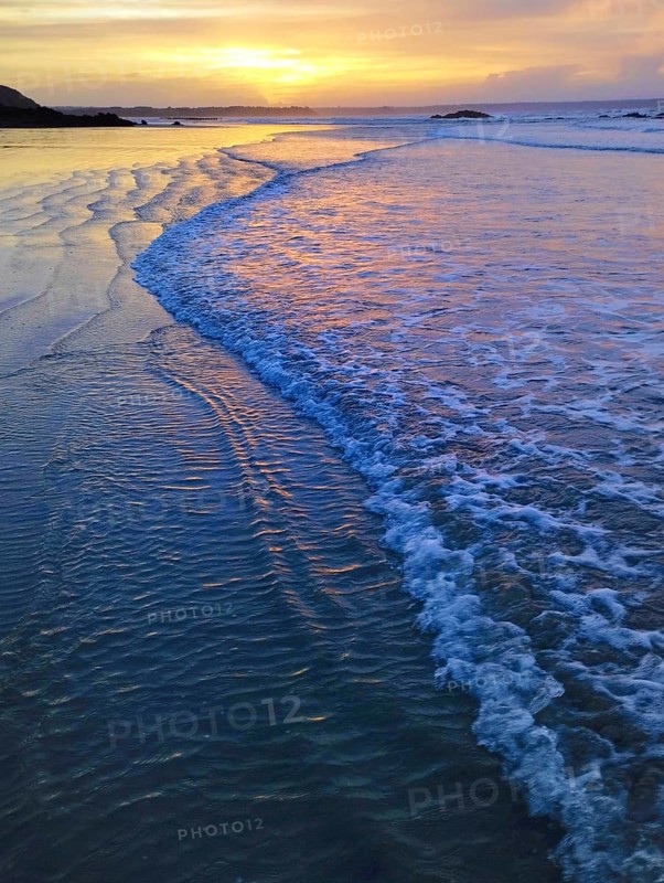 A serene beach scene with gentle waves reflecting a vivid sunset sky, creating a calm and peaceful atmosphere. Planguenoual