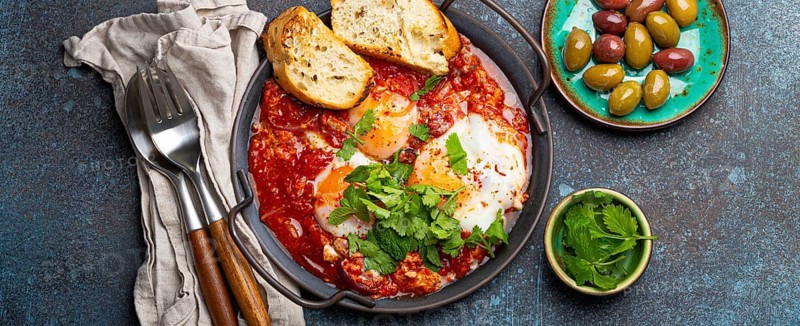 Middle Eastern and Maghrebi healthy dish Shakshouka made of eggs and tomato sauce served in pan with toasts, fresh cilantro and olives on rustic concrete background table from above, food photography