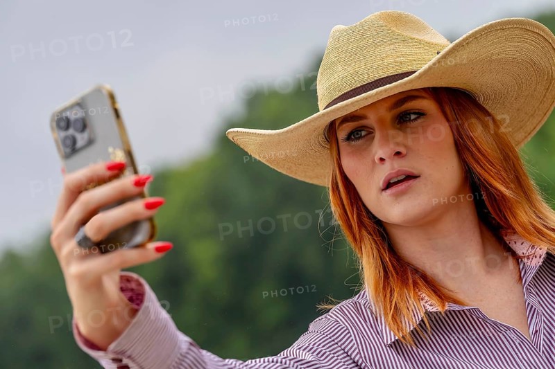 A beautiful redhead model uses her cellphone while posing in a country setting wearing cowgirl attire