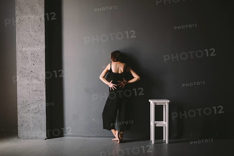 Social theme of female loneliness is pain suffering. Abastration of male domestic violence. Young beautiful Caucasian woman in black clothes, strange pose, white chair big dark room. Fear and despair