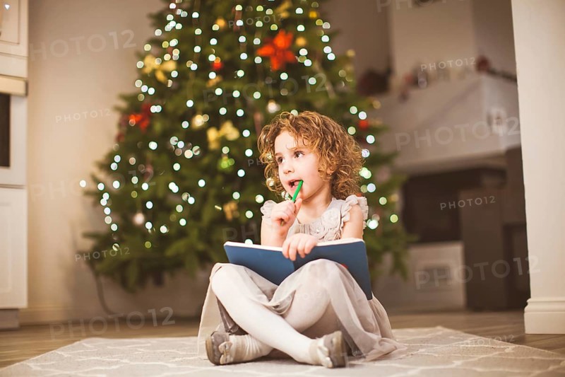 Sweet dreamy little girl with curly hair, wearing grey dress, sitting in front of decorated Christmas tree, with her legs crossed. Girl thinking about letter to Santa, holding notebook and pencil