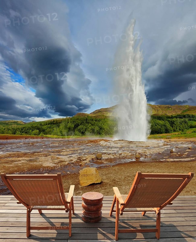 Pillar of hot water and steam from forcing its way out of the ground. Geyser Strokkur in Iceland. Two lounge chairs and small table on wooden platform for easy observation