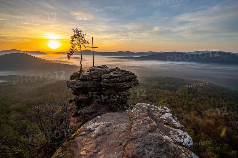 Sunrise on a rock with a summit cross and a view over a forest. Morning atmosphere with fog in the middle of nature. Landscape in the Palatinate Forest, Germany, Europe