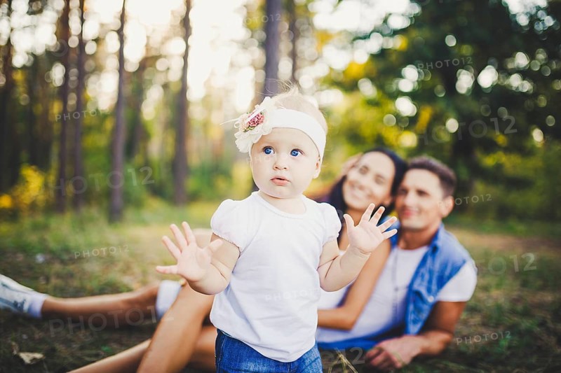 Little girl one year on the background of parents resting on graß learning to walk on nature in the park. The first steps of a child in nature outside the city in the forest