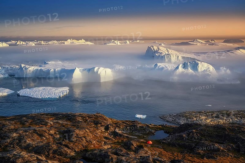 Large glaciers in the bay by the rocky shore in the light of the setting sun with fog over the water with orange tourist tent