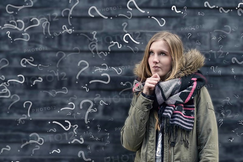 Pretty young female student in class with a problem to solve standing in front of a chalkboard in class covered in question marks looking thoughtfully into the air with her hand to her chin