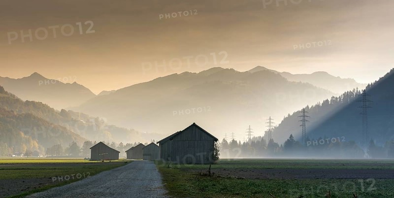View of the Praettigau Valley near Klosters in Switzerland on an early morning with farm fields and wooden barns and power lines and mountains in silhouette behind in late autumn