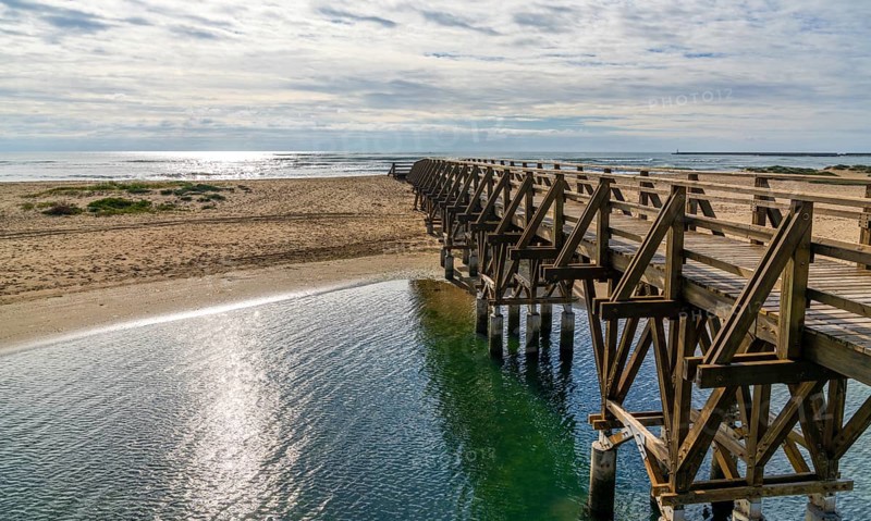 A long wooden boardwalk pier leads from one beach to another over a small ocean inlet