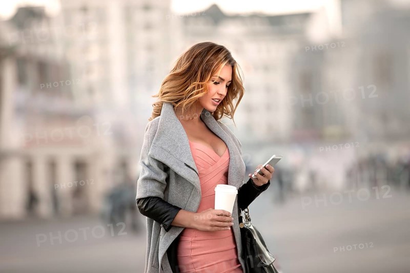A businesswoman checking emails on her mobile phone and holding a coffee cup in her hand in front of an urban backdrop