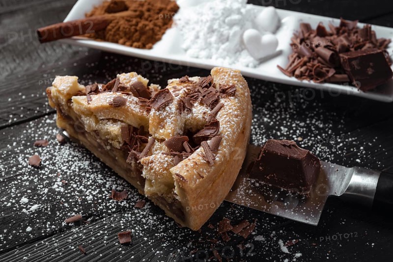 Slice of apple pie with powdered sugar and chocolate, on a knife, on a black rustic table and a plate with cinnamon.Sweet pastry. Baked desserts