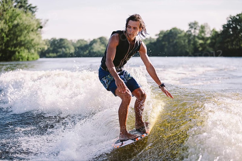 Young athletic man with long hair wakesurfing on waves of river in sunny summer weather. Ttheme outdoor activities in summer. Water sports wakesurf on the board. sliding wakeboarder in water splash
