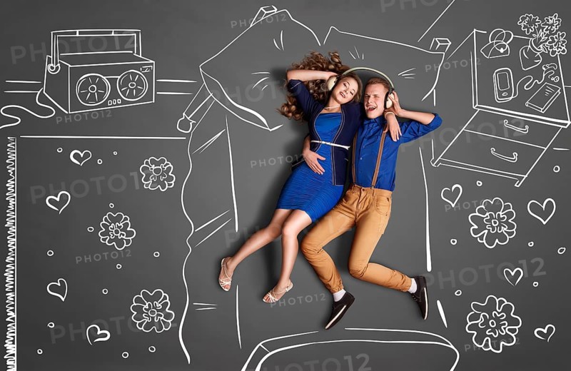 Happy valentines love story concept of a romantic couple lying in bed, sharing headphones, and listening to music against chalk drawings room background