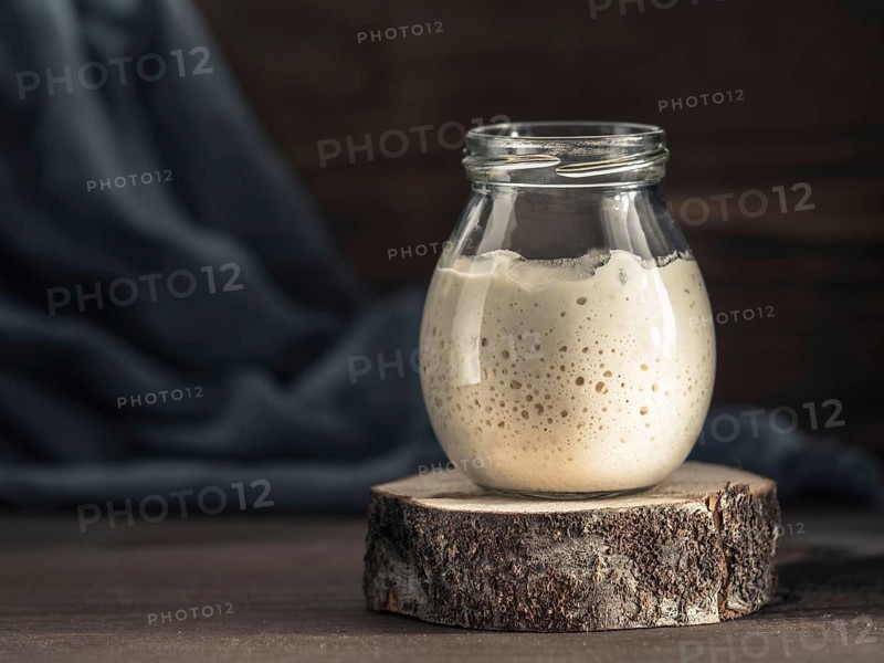 Active wheat sourdough starter in glass jar on brown wooden background. Starter for sourdough bread. Toned image. Copy space