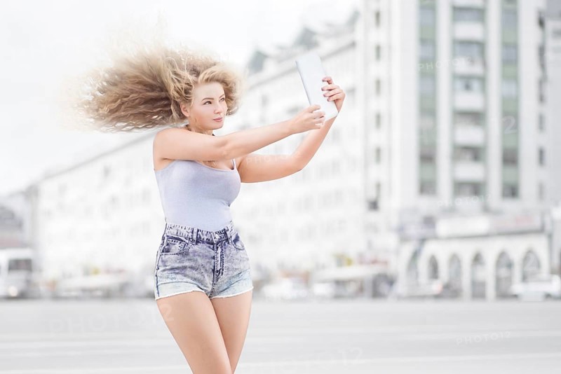 Happy young woman with vintage music headphones around neck taking selfie with tablet PC and smiling happily against urban city background