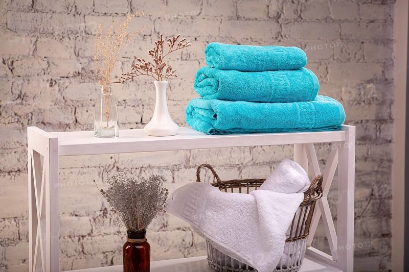 Rack with a stack of three colour turquoise towels and baskets with clean white towels and toilet decor near brick wall. Shelf with terry towels and decorative vases. Shelving unit with clean towels