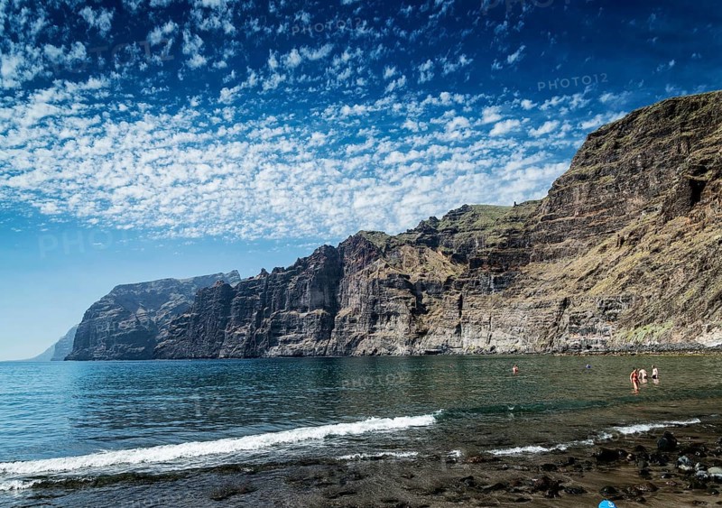 Tourists bathing at los gigantes volcanic black sand beach by famous natural cliffs landmark in south tenerife island spain